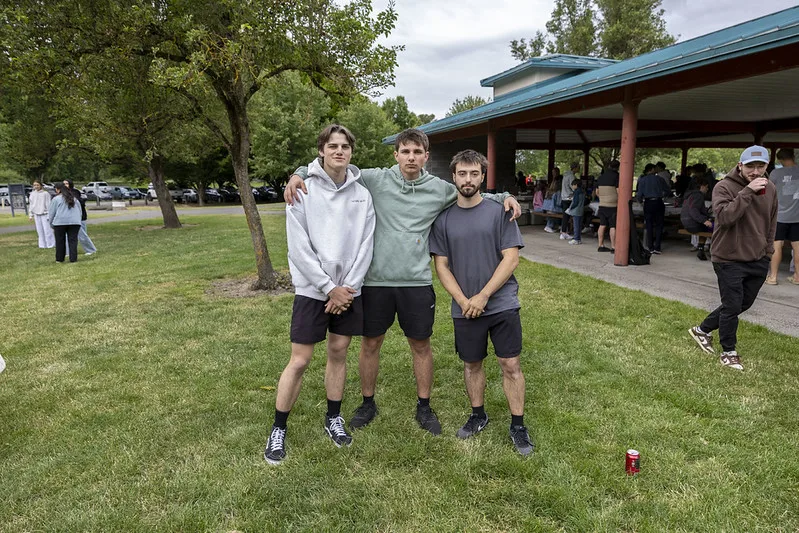Three friends arm in arm at a park pavilion during the youth outing, the rest of the group behind them